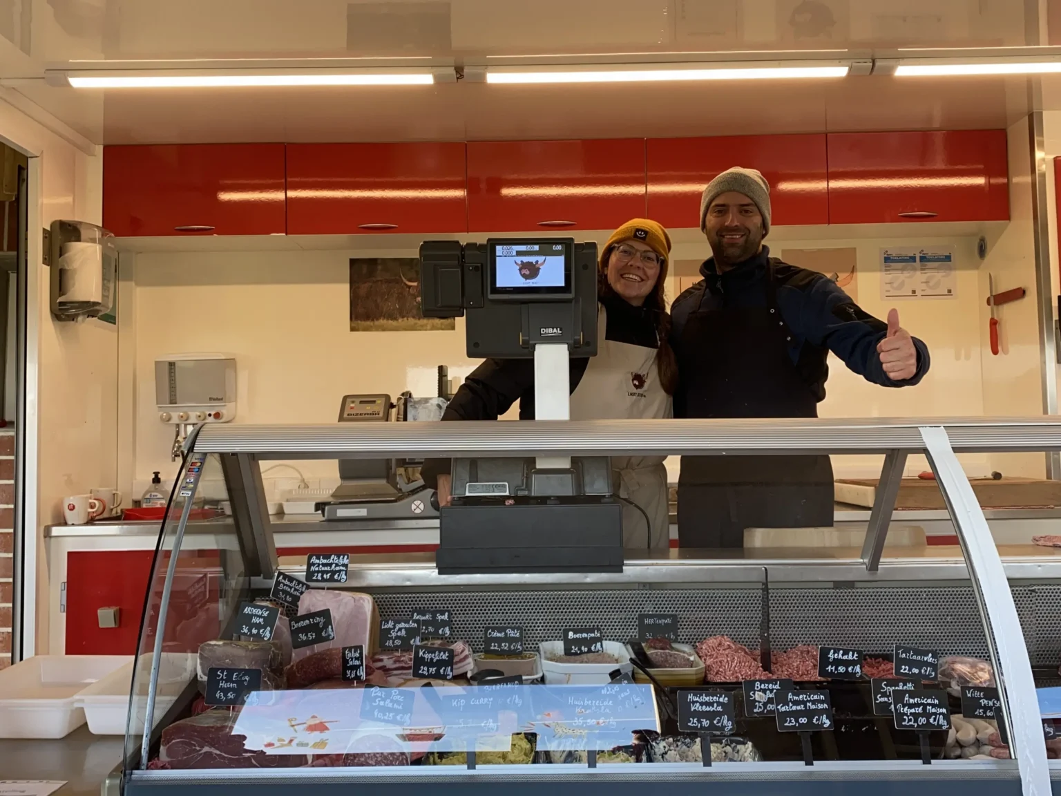 Jeroen en Caro van Lucky Meat glimlachend achter de toonbank van hun hoeveslagerij in Huldenberg, gevuld met ambachtelijke vleeswaren zoals Ardense ham en vers gehakt.